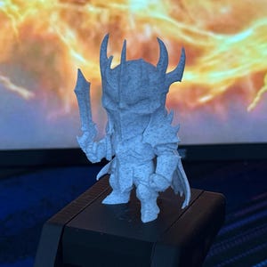 May include: A light gray 3D-printed figurine of a fantasy warrior, holding a sword and wearing armor. The figure has a horned helmet and a cape. The background is a blurred image of a fiery, orange and yellow scene.