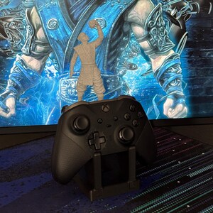Może przedstawiać: Czarny kontroler Xbox spoczywający na czarnym stojaku, z małą szarą figurką na g&oacute;rze. Tło przedstawia niebiesko-białą cyfrową ekspozycję artystyczną. Kontroler ma krzyżakowy d-pad i wiele przycisk&oacute;w.