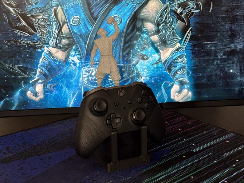 Może przedstawiać: Czarny kontroler Xbox spoczywający na czarnym stojaku, z małą szarą figurką na g&oacute;rze. Tło przedstawia niebiesko-białą cyfrową ekspozycję artystyczną. Kontroler ma krzyżakowy d-pad i wiele przycisk&oacute;w.