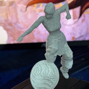 May include: A light gray figurine of a person in a dynamic pose, balanced on a circular base with a water symbol. The figure is detailed with textured surfaces, suggesting a 3D-printed design. The background is blurred.