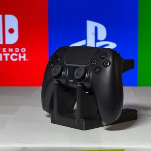 May include: A black video game controller is displayed on a stand in front of a red, blue, and green background. The background features the logos for Nintendo Switch, PlayStation, and Xbox.