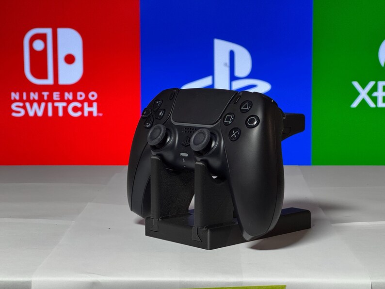 May include: A black video game controller is displayed on a stand in front of a red, blue, and green background. The background features the logos for Nintendo Switch, PlayStation, and Xbox.