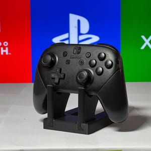 May include: A black Nintendo Switch Pro Controller is standing upright on a black stand. The controller is in front of a red, blue, and green background with the logos for Nintendo Switch, Playstation, and Xbox.