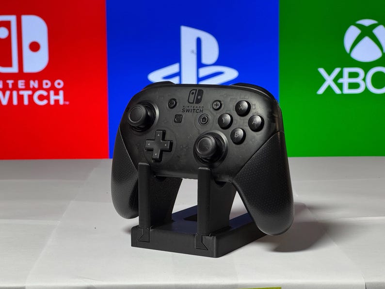 May include: A black Nintendo Switch Pro Controller is standing upright on a black stand. The controller is in front of a red, blue, and green background with the logos for Nintendo Switch, Playstation, and Xbox.