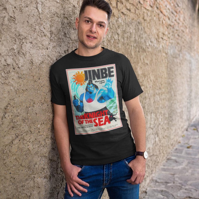 May include: Black t-shirt featuring a graphic of a blue character with the text "JINBE" and "THE KNIGHT OF THE SEA". The graphic has a vintage poster style with red, white, and blue accents. The shirt is being worn by a person.