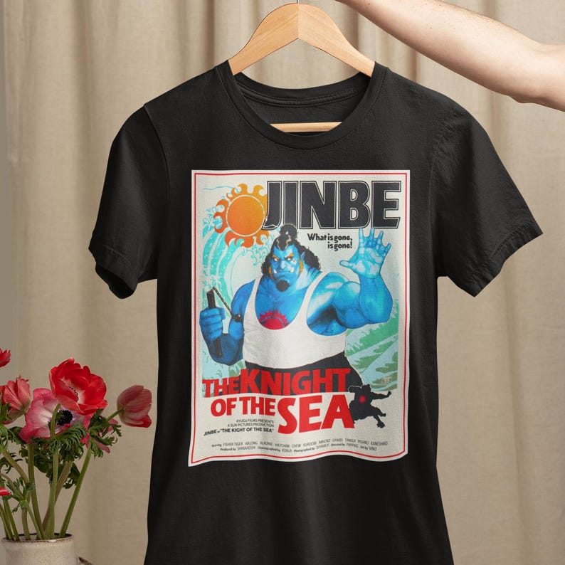 May include: Black t-shirt featuring a graphic of a blue character with the text "JINBE" and "THE KNIGHT OF THE SEA". The design includes a sun and the phrase "What is gone, is gone!" The shirt is hanging on a wooden hanger.