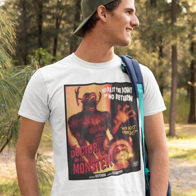 May include: White t-shirt featuring a vintage movie poster design. The poster has a dark red and brown color scheme with the text "THE DOCTOR AND THE MONSTER" and "HE'S PAST THE POINT OF NO RETURN". The design also includes a monster and a small character.