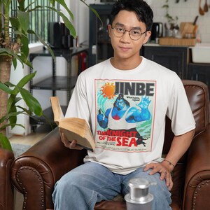 May include: A person wearing a white t-shirt with a graphic of a muscular blue character, the text "JINBE", and "THE KNIGHT OF THE SEA". The person is sitting in a brown leather chair and holding a book.