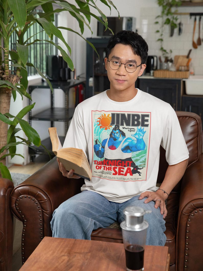 May include: A person wearing a white t-shirt with a graphic of a muscular blue character, the text "JINBE", and "THE KNIGHT OF THE SEA". The person is sitting in a brown leather chair and holding a book.