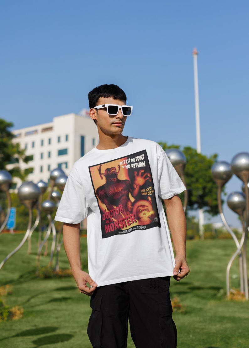 May include: White t-shirt featuring a vintage movie poster design for "Doctor and the Monster." The poster art includes a monster illustration and text. The shirt is worn outdoors on a sunny day.