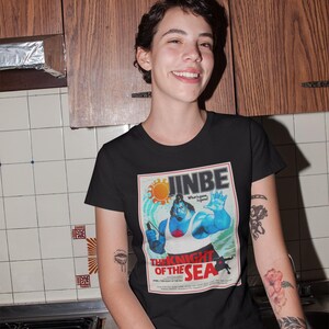 May include: Black t-shirt featuring a vintage-style graphic of a blue character with the text "JINBE" and "THE KNIGHT OF THE SEA." The design includes a sun and a figure holding a weapon. The shirt is made of a soft material.