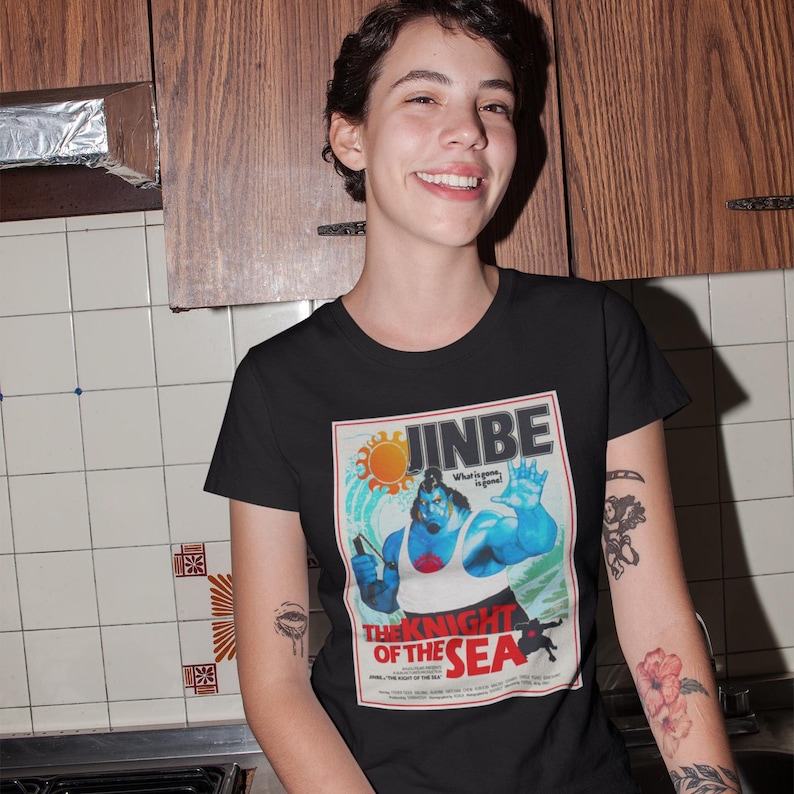 May include: Black t-shirt featuring a vintage-style graphic of a blue character with the text "JINBE" and "THE KNIGHT OF THE SEA." The design includes a sun and a figure holding a weapon. The shirt is made of a soft material.
