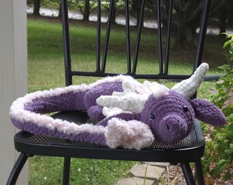 May include: A handmade crochet dragon in shades of purple, white, and pink rests on a black metal chair. The dragon features a long, plush tail and detailed facial features, creating a whimsical and cuddly design.