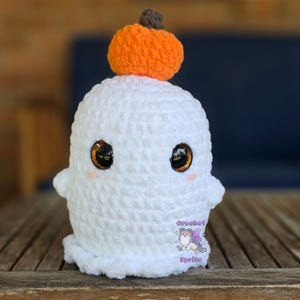 May include: A handmade white crochet ghost with large, expressive eyes and a small orange pumpkin on top. The ghost has small arms and a cute, friendly expression. The Crochet Sprite logo is visible on the bottom.