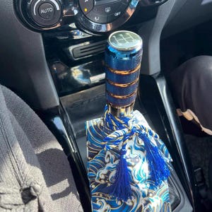 以下が含まれることがあります： 青と金のシフトノブと、青と金の波模様のシフトブーツ。ブーツにはタッセルが付いています。ノブには5速のギアパターンがあります。