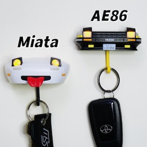 Könnte beinhalten: Zwei schlüsselanhänger in Form von Autos, einer weiß mit einer roten Zunge und dem Text "Miata" darüber, der andere schwarz mit dem Text "AE86" und "TRUENO" darüber. Beide Haken haben gelbe Scheinwerfer und sind an Schlüsselringen befestigt.