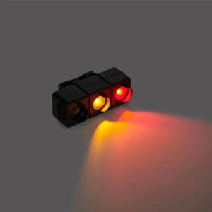 Op de afbeelding: Een kleine, zwarte verkeerslicht met drie verlichte secties: groen, geel en rood. De gele en rode lichten zenden licht uit. Het apparaat heeft een clip en is waarschijnlijk een veiligheids- of signaalapparaat.