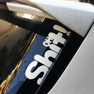 May include: A white vinyl sticker with the text "Oh Shit!" on a black background, attached to the back window of a silver car.