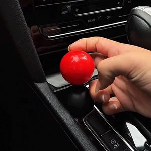 Może przedstawiać: Gałka zmiany biegów w kształcie czerwonej kuli z czarną podstawą. Gałka jest trzymana przez rękę w samochodzie.