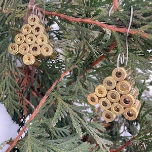 May include: Two Christmas tree ornaments made from brass bullet casings. The ornaments are arranged in a triangular shape and suspended by silver wire hooks. The background features green evergreen branches.