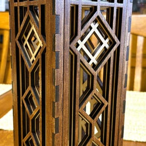 Pode incluir: Uma lanterna decorativa de madeira com um design geométrico. A lanterna é retangular com formas de diamante e angulares recortadas. A madeira tem um tom marrom quente com detalhes em preto. A lanterna é provavelmente para decoração de casa.