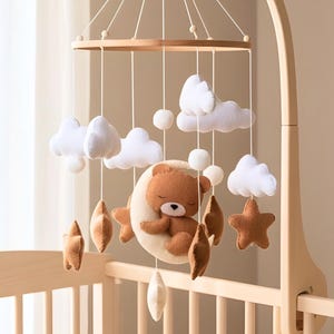 Pode incluir: Móvel de berço com um ursinho de pelúcia castanho dormindo em uma lua crescente creme, rodeado por nuvens brancas, estrelas castanhas e esferas creme. O móvel é suspenso por uma estrutura de madeira.