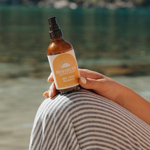 Op de afbeelding: Een bruine fles SPF 15 shimmer spray met een wit etiket met een geel zonontwerp en de tekst "Sunshine by Sun Milk". De fles wordt vastgehouden door een persoon die een gestreepte broek draagt.