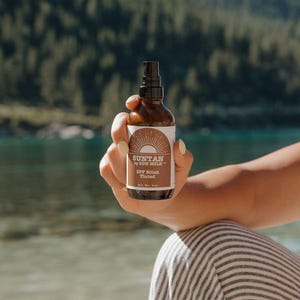 Könnte beinhalten: Eine braune Flasche Suntan by Sun Milk SPF 30ish Tinted Sonnencreme wird vor einer malerischen Kulisse aus See und Bäumen gehalten. Das Etikett zeigt ein Sonnenstrahl-Design und den Produktnamen.