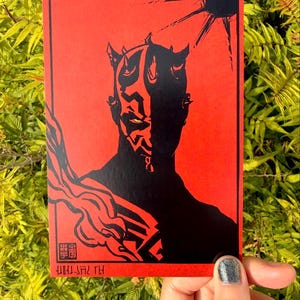 Può includere: Una stampa artistica rettangolare che presenta la sagoma nera ad alto contrasto di un personaggio cornuto con occhi gialli su uno sfondo rosso vibrante. Il design include fumo nero stilizzato, un sole e del testo rosso in basso.