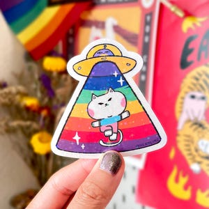 May include: A die-cut sticker featuring a white cat with pink cheeks wearing a blue and pink shirt, being abducted by a yellow UFO. The beam is a rainbow gradient with a holographic star pattern. The text HERBLINZ is printed in the bottom right corner.