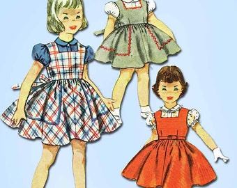 Op de afbeelding: Vintage naaipatroon voor meisjesjurken. Het patroon toont drie meisjes die verschillende stijlen jurken dragen. De eerste jurk is geruit, de tweede is grijs en de derde is rood. Het patroon is van Vintage4me2.