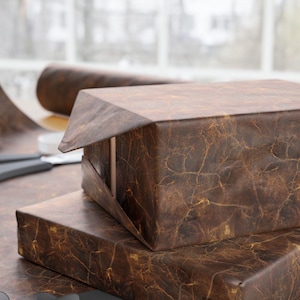May include: Two brown gift boxes wrapped in textured paper with a rustic look. The top box is open, revealing a brown interior. The boxes are sitting on a brown surface with a black ribbon.