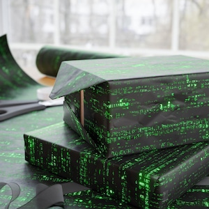 May include: A stack of three gift boxes wrapped in black and green wrapping paper with a pattern of green code. The boxes are sitting on a table covered in the same wrapping paper.