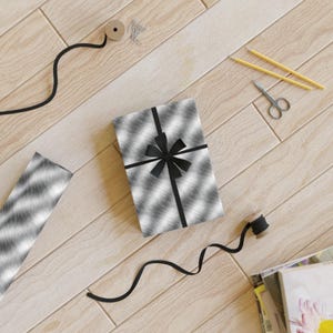 May include: A black and white patterned gift box with a black ribbon and bow. The box is sitting on a wooden surface with black ribbon, a spool of ribbon, and pencils.
