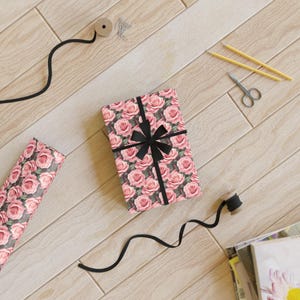 May include: A gift box wrapped in pink floral patterned paper with a black ribbon bow. The box is on a wooden surface with other craft supplies.