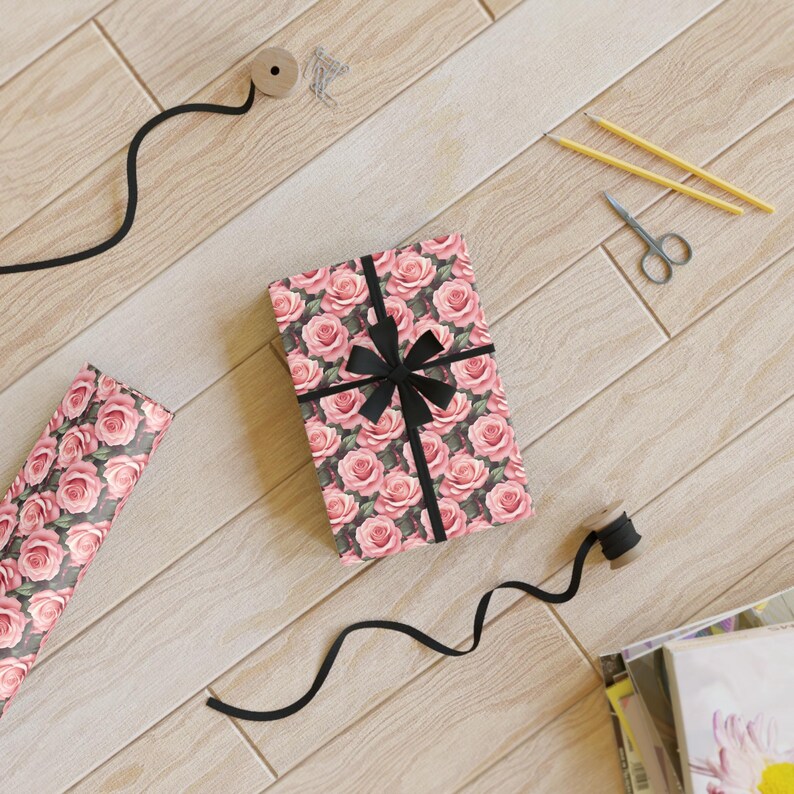 May include: A gift box wrapped in pink floral patterned paper with a black ribbon bow. The box is on a wooden surface with other craft supplies.