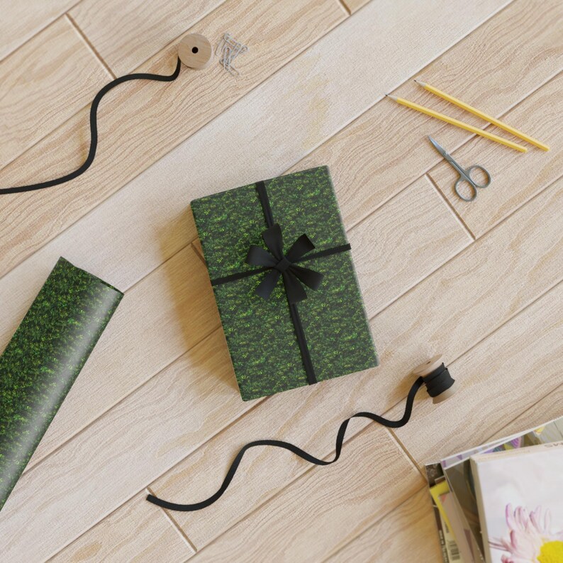 May include: A green and black gift box with a black ribbon and bow. The box is surrounded by wrapping paper, ribbon, scissors, pencils, and a spool of thread.