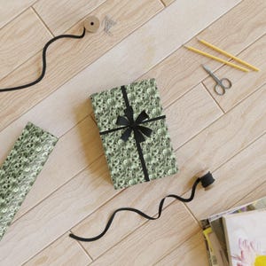 Op de afbeelding: Een groene en zwarte cadeaupapier met een schedelpatroon. Het cadeaupapier is om een ​​doos gewikkeld met een zwart lint en strik. De doos staat op een houten oppervlak.