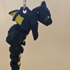 May include: A black and yellow dragon keychain. The dragon has yellow accents on its wings and tail. The keychain is made of a 3D-printed material and is attached to a silver metal clasp.