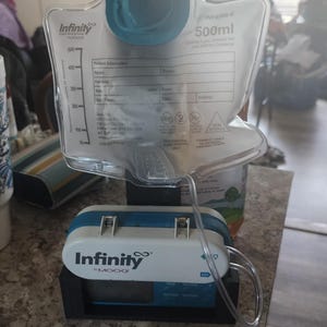 May include: A medical feeding system featuring a clear plastic bag with a 500ml capacity, connected to a white and blue pump labeled "Infinity". The system is designed for medical use and is placed on a countertop.