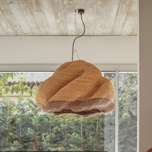 May include: A woven pendant light with a distinctive, textured design. The light is a warm tan colour with a darker brown trim at the bottom. It hangs from a black cord and is suspended from a concrete ceiling. The light is positioned in front of a window with a view of green trees.