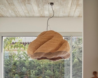 May include: A woven pendant light fixture with a unique, textured design. The light is a warm tan color with a darker brown trim at the bottom. It hangs from a black cord and is suspended from a concrete ceiling. The light is positioned in front of a window with a view of green trees.