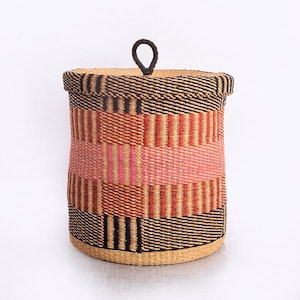 May include: A round woven storage basket with a lid and a handle. The basket is made of natural fibres and features a mix of colours, including black, brown, pink, and tan. The design incorporates horizontal and vertical stripes. The handle is a small, dark loop.