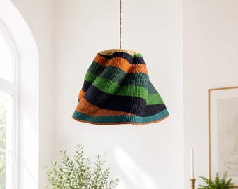 Pode incluir: Uma luminária pendente com uma cúpula tecida em forma de sino com listras horizontais em tons de laranja, azul escuro e verde. A cúpula está suspensa por um cordão fino e trançado marrom que se estende para cima a partir do centro.