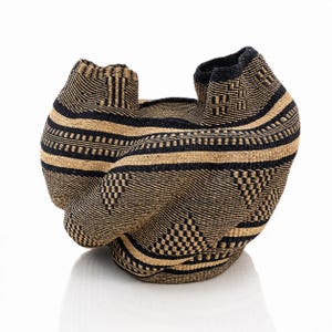 May include: A woven basket with a flexible, organic shape featuring a repeating geometric pattern in black and natural tan fibers. The structure has an asymmetrical, folded rim and a dense, intricate weave throughout its entire exterior surface.