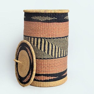 Può includere: Un cesto alto e cilindrico intrecciato con coperchio. Il cesto presenta fasce alternate di motivi intrecciati marrone rossiccio e neri. Il coperchio è rotondo con una maniglia e cerchi concentrici neri e marroni. Il cesto viene probabilmente utilizzato per riporre oggetti o per il bucato.