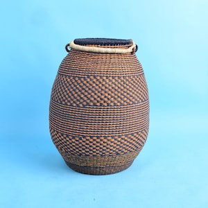 Op de afbeelding: Een geweven mand met een afgeronde body en een donkerblauw en bruin geometrisch patroon. De rand is donkerblauw met een lichtbruine rand. Twee kleine handvatten zitten aan de zijkanten van de rand. De mand staat tegen een lichtblauwe achtergrond.