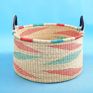May include: A round woven basket with red and teal diagonal stripes. The rim is trimmed with red and teal, and it has two black and white handles. The basket is made of natural-coloured woven material.