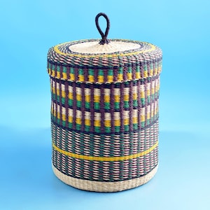 May include: A cylindrical woven basket with a lid. The basket features a pattern of green, yellow, and purple horizontal stripes. The lid has a small, dark handle. The basket is approximately 30 cm tall and 20 cm in diameter.