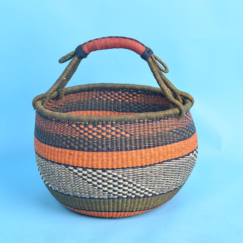 May include: A handwoven basket with a rounded shape and a sturdy handle. The basket features horizontal bands of orange, black, white, and green. The handle is wrapped in red leather. This decorative basket is ideal for storage or display.